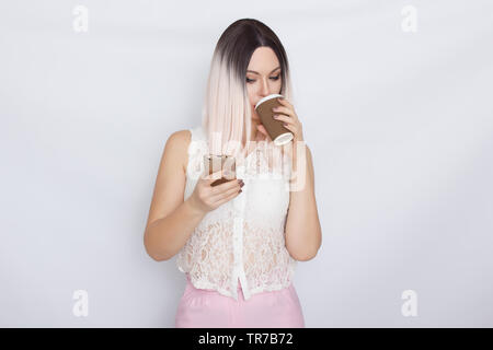 Image de jolie belle jeune femme blonde en chemise blanche posant sur fond blanc à boire du café et à l'aide de son shartphone Banque D'Images