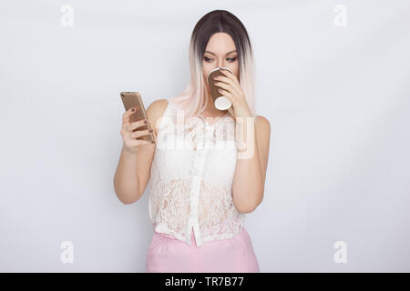 Image de jolie belle jeune femme blonde en chemise blanche posant sur fond blanc à boire du café et à l'aide de son shartphone Banque D'Images