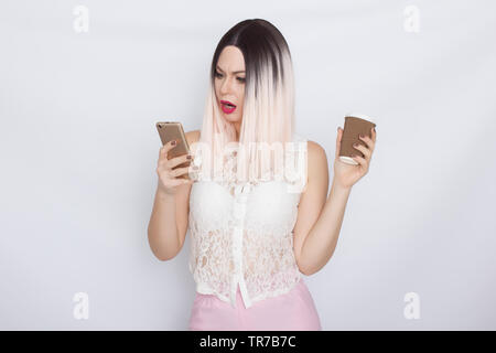 Image de jolie belle jeune femme blonde en chemise blanche posant sur fond blanc à boire du café et à l'aide de son shartphone Banque D'Images