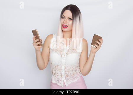 Image de jolie belle jeune femme blonde en chemise blanche posant sur fond blanc à boire du café et à l'aide de son shartphone Banque D'Images