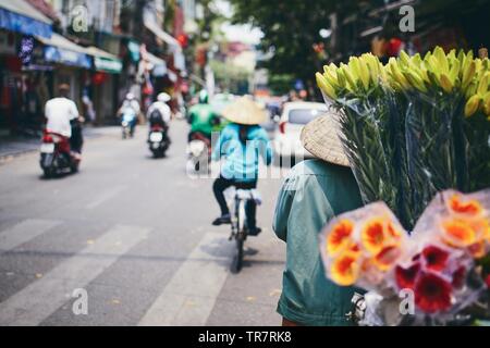 La vie en ville dans la rue de vieux quartier de Hanoi. Vendeuse de fleurs dans un chapeau conique, Vietnam. Banque D'Images