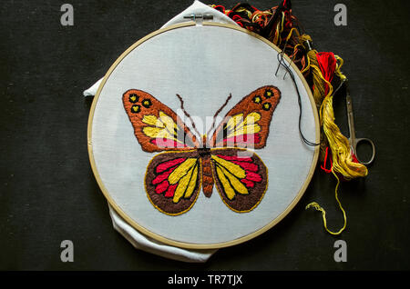 Papillon lumineux stylisé brodé sur tissu de coton blanc, tendue sur un cercle en bois avec différentes couleurs de fils et ciseaux couché sur contreplaqué noir Banque D'Images