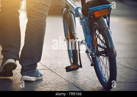 Un homme habillé avec un jean bleu et des chaussures de marche sur le trottoir, éclairé par la lumière du soleil, et porte un bike bleu Banque D'Images