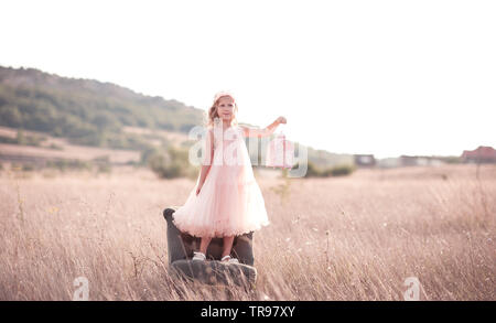 Cute kid fille 5-6 ans rose dress posing wearing élégant avec une chaise dans le pré. À la voiture. Banque D'Images