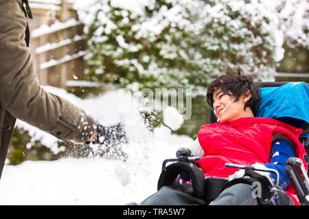 Mobilité biracial mignon garçon âgé de douze ans en fauteuil roulant assis dehors en hiver, jouer avec un adulte throwing snow and smiling Banque D'Images