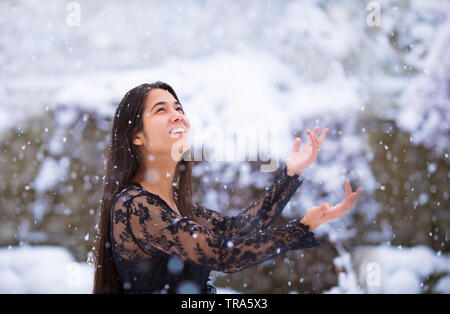 Smiling girl biracial robe dentelle noire à l'extérieur en hiver attraper des flocons en main tendue Banque D'Images