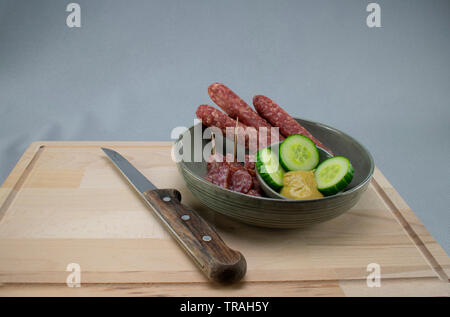 Petit bol avec saucisse de porc tranches de cornichon et de la moutarde sur une planche en bois avec un couteau bien aiguisé et un fond gris Banque D'Images