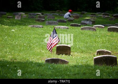 Plymouth Meeting, PA, USA - 1 juin 2019 : A sunlit drapeau américain se pare d'une tombe de premier plan tandis qu'au loin un jeune garçon, en bonnet rouge s'agenouille Banque D'Images