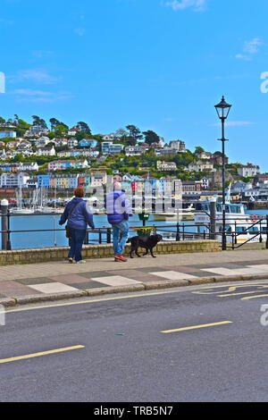 Un couple en train de marcher avec leur chien le long du front de mer de Dartmouth donnant sur la rivière Dart vers le pittoresque village perché de Kingswear. Banque D'Images