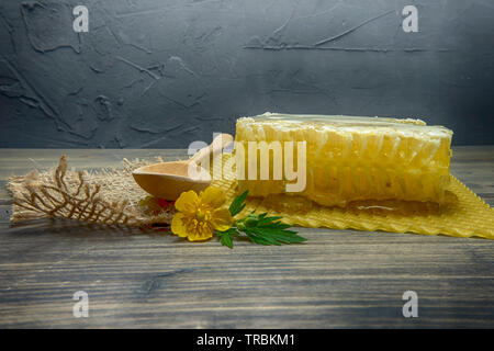 Bloc de rayons à miel frais avec du miel qui coulait sur le tapis en dessous avec une cuillère en bois et coloré de fleurs d'onagre jaune, copy space sur bac gris Banque D'Images