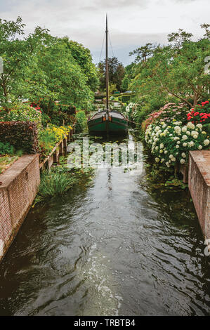 Charmant petit canal avec bateau décoratif à côté et luxuriant jardin fleuri à Drimmelen. Un petit hameau avec port et élégantes rues en Pays-Bas. Banque D'Images