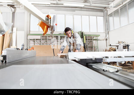 Carpenter l'homme travailler dans la menuiserie, couper une planche de bois avec une scie circulaire, la machine est protégé par un cache-oreilles et des lunettes Banque D'Images