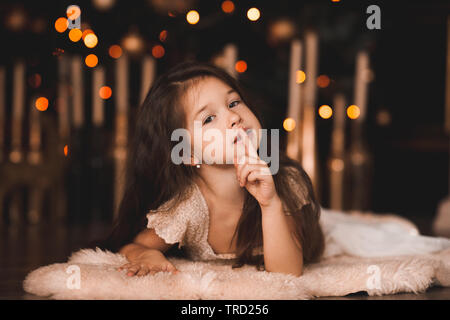 Pretty baby girl making hush allongé sur flor sur les lumières de Noël dans la chambre. En regardant la caméra. Saison de vacances. Banque D'Images