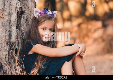 Smiling girl enfant 5-6 ans portant bandeau unicorn et robe noire assis sous arbre en parc. En regardant la caméra. L'enfance. Banque D'Images