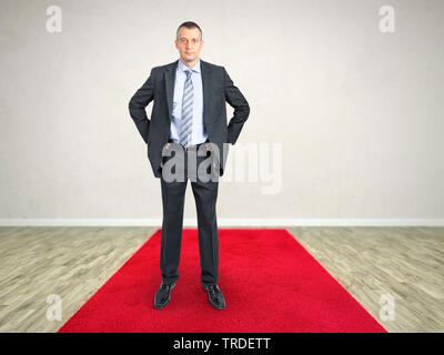 Portrait d'un homme dans un costume gris debout sur un tapis rouge avec les mains dans ses mains sur les hanches Banque D'Images