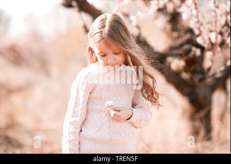 Cute blonde jeune fille 5-6 ans debout à l'extérieur. Holding flower sur fond nature. Le port de chandail rose élégant. L'enfance. Banque D'Images