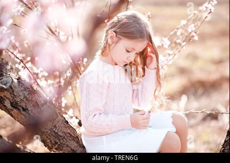 Cute baby girl sitting on tree jouant avec des fleurs à l'extérieur. L'enfance. Banque D'Images