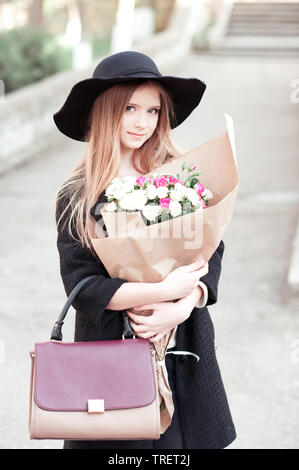 Smiling teen girl 14-16 ans portant veste hiver et hat, holding sac élégant et roses enveloppé dans du papier craft à l'extérieur. En regardant la caméra. Beau Banque D'Images