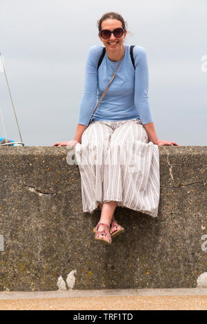 Femme au début 40 s / 40 s / 40 assis sur le mur et sourit dans le soleil chaud. Elle est en jupe, sandales et lunettes de soleil pour protéger du soleil. England UK (99) Banque D'Images