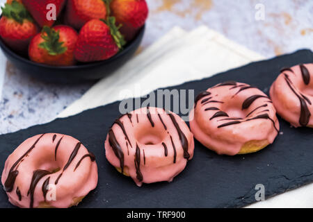 Strawberry-Glazed Donuts avec un filet de chocolat noir sur conseil de béton avec des fraises fraîches dans l'arrière-plan Banque D'Images