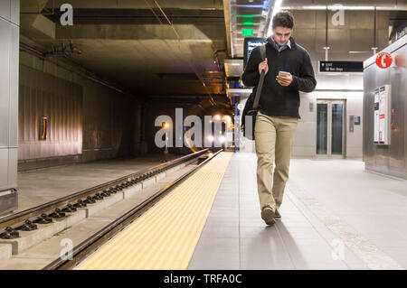 Homme professionnel commuter avec cell phone marche sur la plate-forme de la station de métro comme un train s'approche de la consultation des messages ou en utilisant une application mobile sur wifi Banque D'Images