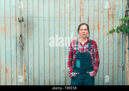 Portrait of female farmer belle ferme en face du hangar. Certain travailleur agricole portant des jeans et chemise à carreaux avec bretelles est à la recherche d'un Banque D'Images