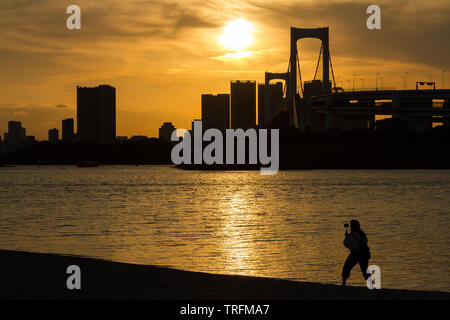 Une femme, en silhouette, utilise un bâton de selfie pour prendre une photo de la vue de Rainbow Bridge depuis une plage à Odaiba, Tokyo, Japon. Banque D'Images