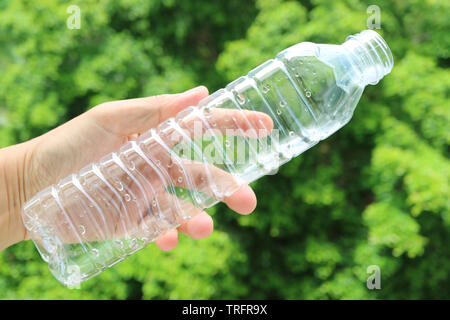 La main de l'homme tenant une bouteille en plastique vide de l'eau potable contre le feuillage vert floue Banque D'Images