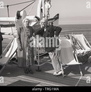 Années 1950, historique, un couple de personnes âgées sur une croisière sur le pont extérieur permanent de la P & O transatlantique SS Chusan. La dame porte une robe longue, avec un manteau, tandis que l'homme n'est plus convenablement vêtue pour une croisière dans un blazer et des chaussures de pont et est fumant une pipe. Banque D'Images