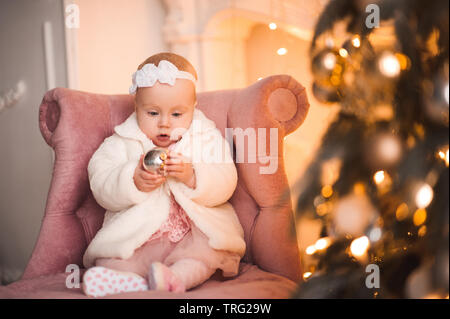 Bébé fille 1 ans holding Christmas ball assis sur chaise élégante rose en vertu de l'arbre de Noël dans la salle. Saison de vacances. Banque D'Images