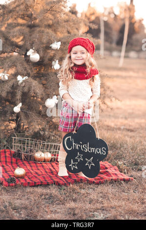 Smiling girl enfant 4-5 ans portant bonnet et écharpe en tricot noël décor extérieur. holding Arbre de Noël avec des décorations. Le temps des vacances. Banque D'Images