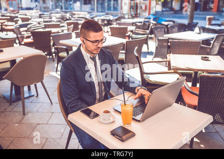 Beau jeune homme assis au café de la rue de la saisie sur ordinateur portable et portant des écouteurs. Banque D'Images