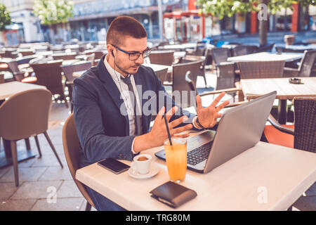 Jeune homme assis au café de la rue d'avoir une conversation sur un ordinateur portable portant un casque sur la tête de parler a l'air en colère levant la main. Banque D'Images