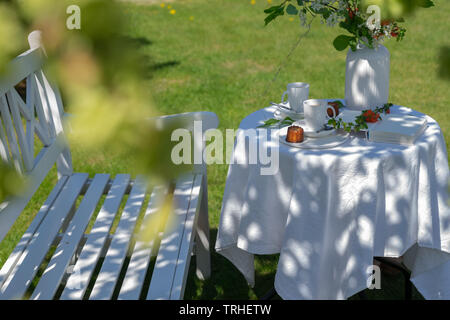 Tableau blanc servi avec café, caneles et fleurs à côté du blanc banc dans le jardin. Profitant de la journée ensoleillée et reposante Banque D'Images