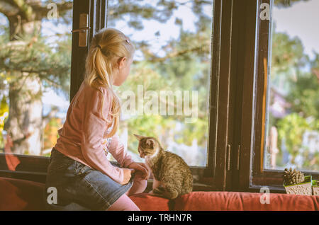 Adorable petite fille blonde avec cat assis à côté de la fenêtre avec vue sur la forêt Banque D'Images