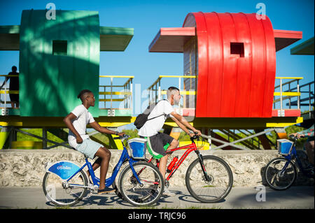 MIAMI - Juillet 2017 : les touristes en utilisant CitiBike système de partage de vélos balade le long de la promenade de South Beach en face d'une rangée de tours de sauveteur colorés. Banque D'Images