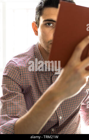 Portrait d'un jeune homme aux cheveux noirs et une chemise à la caméra à tout en lisant un livre. Se concentrer sur l'oeil avec effet de flou, l'arrière-plan lumineux. Banque D'Images