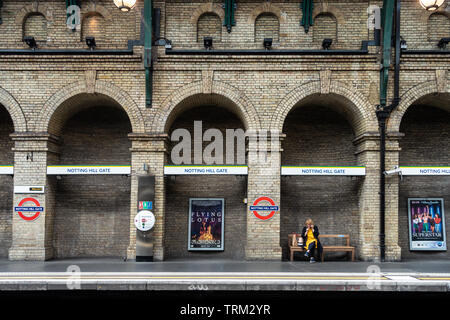 Une dame assise seule, à parler sur son téléphone portable, sur un banc sur une plate-forme tranquille à Notting Hill Gate London Underground Station. Banque D'Images