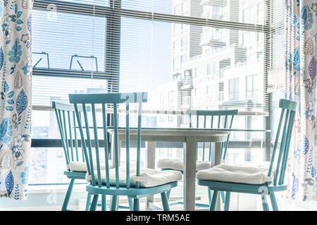 Salle à manger très lumineuse dans un appartement, avec de grandes fenêtres à l'arrière-plan, et le bleu des chaises avec une table en verre à l'avant-plan. Rideaux Banque D'Images