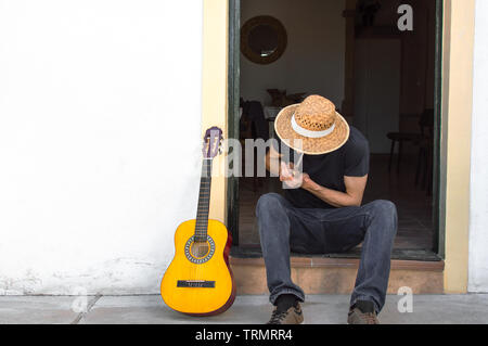 Jeune homme au chapeau de paille et une guitare fumer un joint de marijuana à la porte d'une maison blanche dans la campagne. Le guitariste assis dans la rue. Banque D'Images