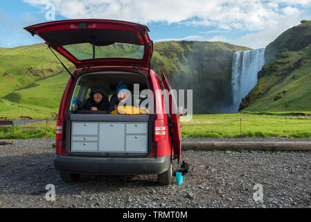 Couple dans une fourgonnette rouge dans des sacs de couchage avec porte arrière ouverte. Skogafoss chute d'eau dans l'arrière-plan, l'Islande Banque D'Images
