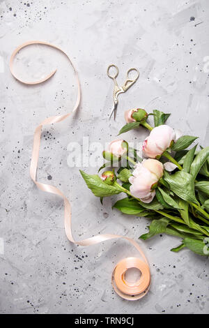 Bourgeons délicats avec des pivoines et des feuilles vertes sur un tableau gris du fleuriste à côté d'un ruban de corail et ciseaux Banque D'Images