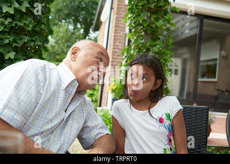 Portrait d'un grand-père et sa jeune petite fille assis dans le jardin ensemble tirant grimaces dans le soleil d'été Banque D'Images