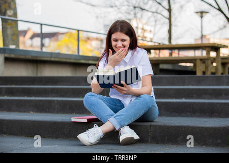 Student girl smiling lire son livre préféré en plein air sur le campus des escaliers. Étudiante agréable et relaxant de lire un livre sur la construction de l'université zone de Banque D'Images