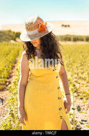 Portrait de jeune femme avec des cheveux noirs bouclés, robe et chapeau de paille jaune avec fleur en place naturelle au coucher du soleil. Femme regarder vers le bas dans la campagne. Banque D'Images