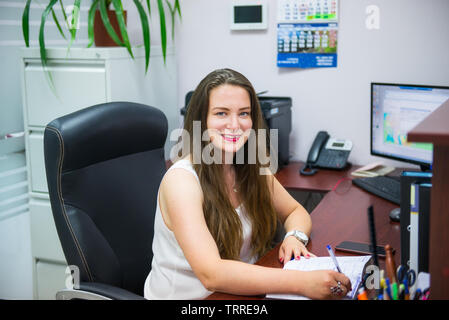 Young caucasian business lady est looking at camera et souriant tout en restant assis sur son lieu de travail en bureau. L'entrepreneuriat féminin. Portrait de main Banque D'Images