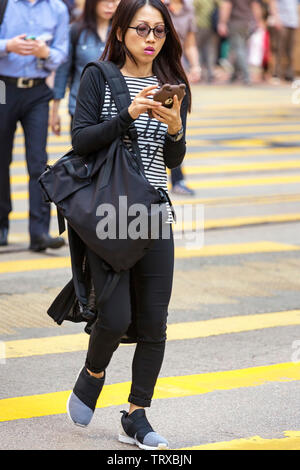 Femme Asiatique se concentrant sur appareil mobile en traversant la route à Hong Kong, SAR, Chine Banque D'Images