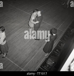 Années 1950, historique, le soir, et deux hommes portant des costumes danser ensemble à une classe de danse, regardé sur le côté par un instructeur féminin, England, UK. Dans cette après-guerre avant de la télévision de masse et de divertissement, des cours du soir d'enseignement comme l'apprentissage de la danse de bal étaient populaires activités sociales pour la population adulte. Banque D'Images