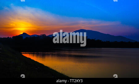 Paysage avec coucher du soleil orange ciel, les silhouettes des montagnes, collines et d'arbres et le lac Banque D'Images