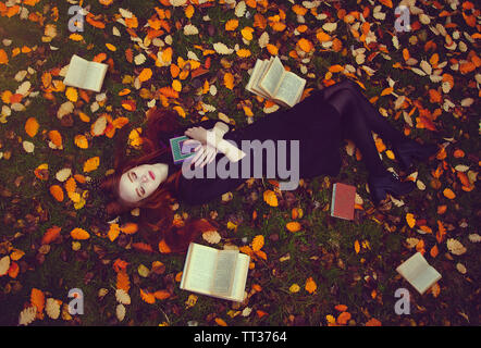 Belle fille aux cheveux roux avec des livres se trouve sur l'herbe dans une forêt d'automne, vue du dessus. Conte d'automne séance photo. Saison d'automne. Concept de confort. Cre Banque D'Images